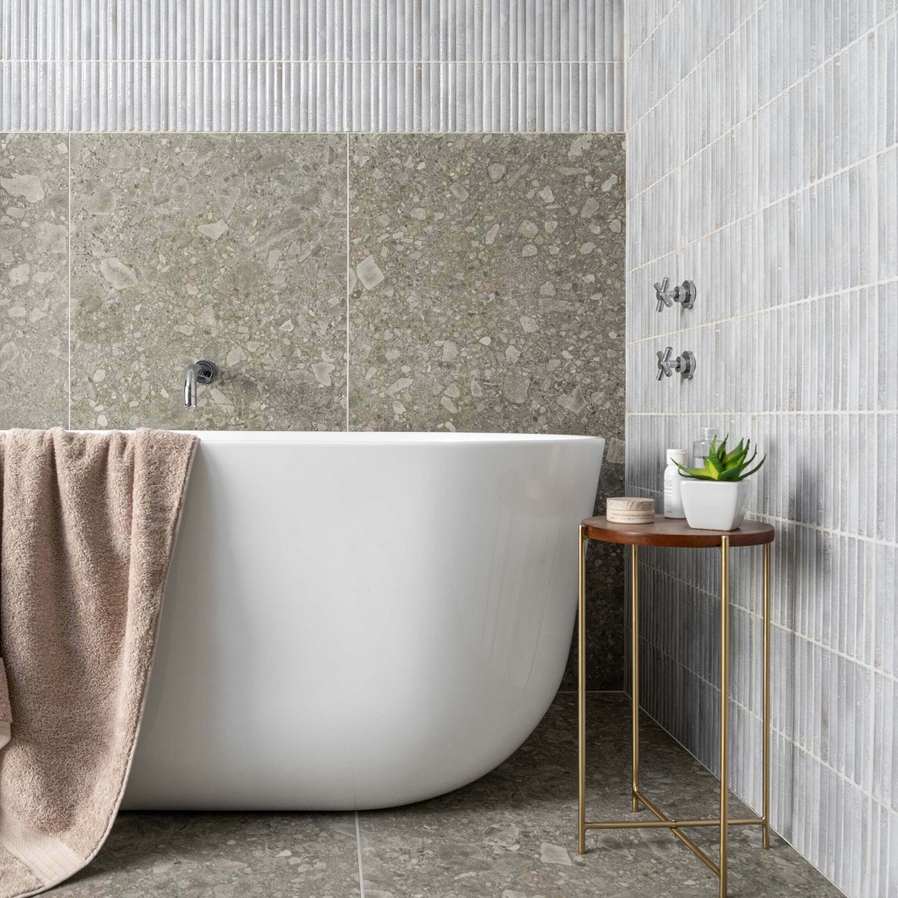 Meraki Kitt-Kat blanco tiles on wall with terrazzo effect tiles on floor and part of wall, styling consisting of towel, silver hardware, and small round table with plant. 