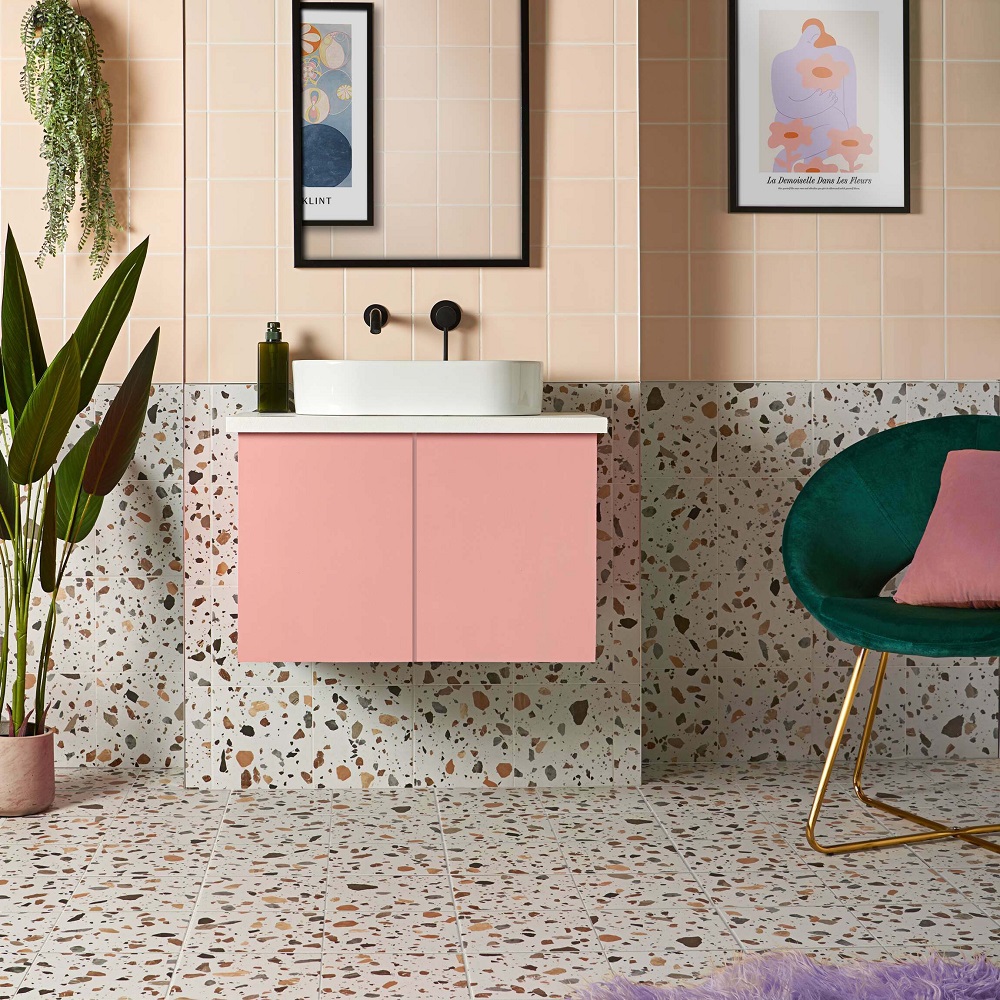 Bathroom colour ideas are reflected in this Playful bathroom with salmon pink square wall tiles, a terrazzo wall and floor. A Peach-pink vanity cabinet with a white basin, black fixtures, framed art, and a touch of greenery from houseplants to complement the aesthetic. 
