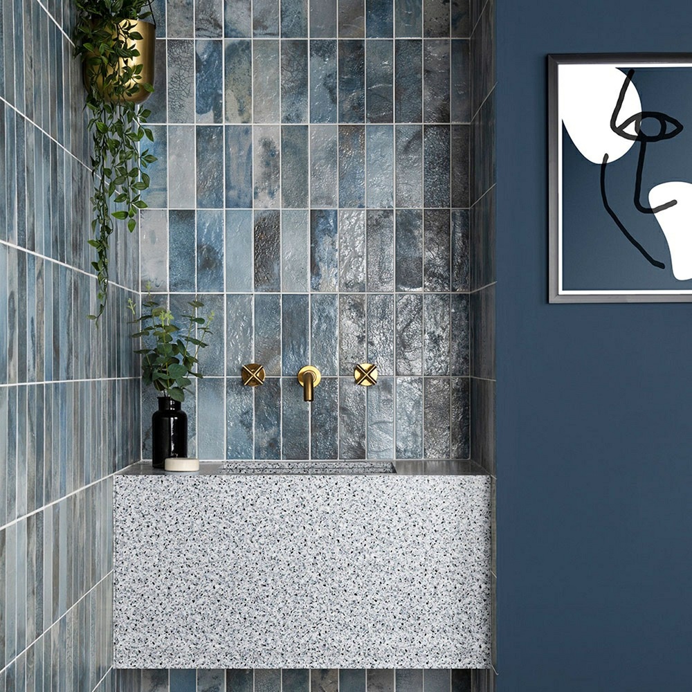 A modern bathroom with a grey terrazzo sink, blue tile splashback, gold taps and hardware for sink, a hanging plant over sink and framed abstract artwork. 
