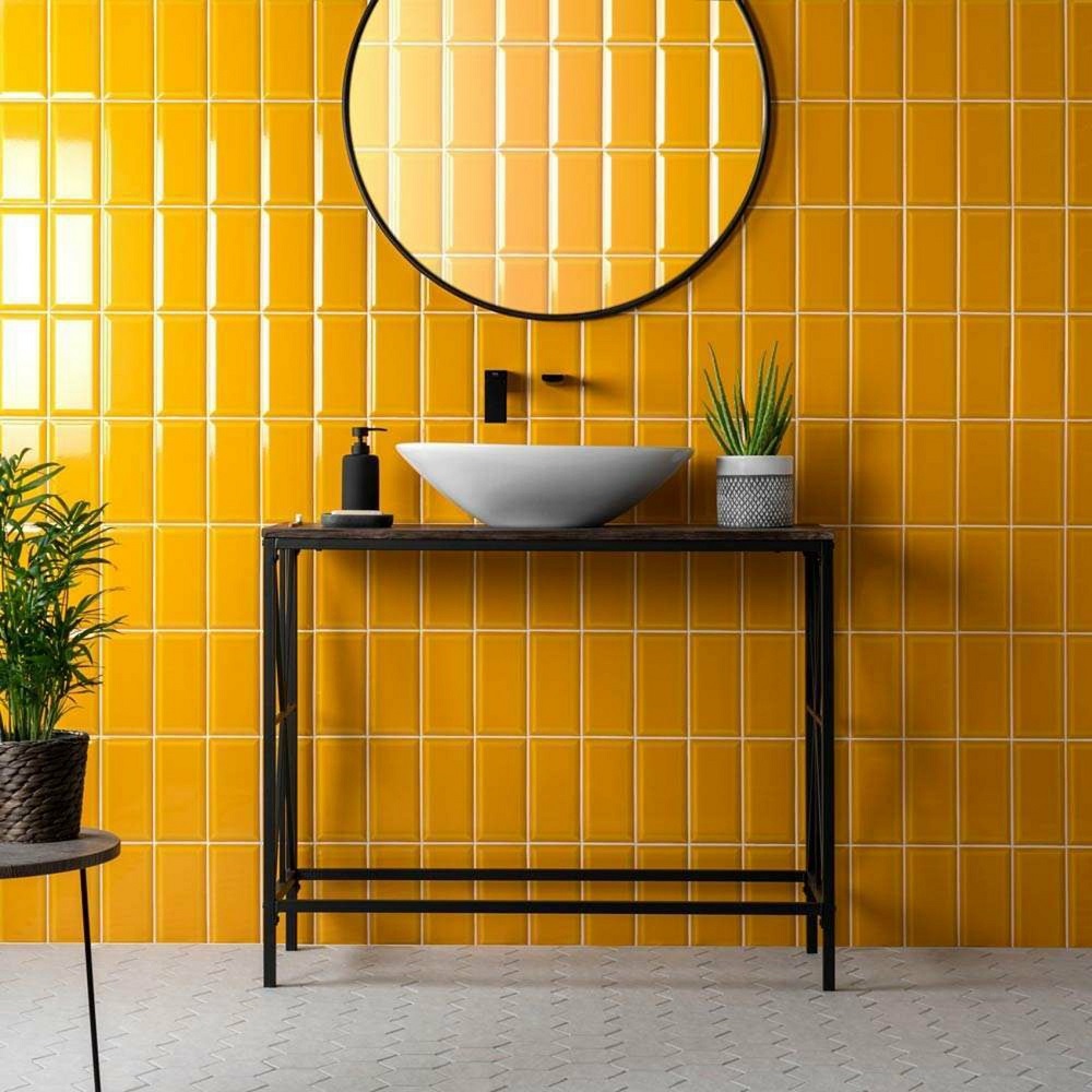 Bathroom wall is vibrant yellow with metro tiles that give an energetic vibe. Sleek round mirror and simple black metal vanity with white vessel sink. Greenery in a couple of pots adds a natural element, and floor is adorned with understated hexagonal tiles, balancing the brightness of the wall. 