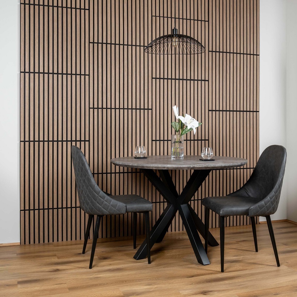 Contemporary dining area with smoked oak square acoustic wall slat panels, round wooden table with cross legs, two elegant grey upholstered chairs, and a striking black wire pendant light. 