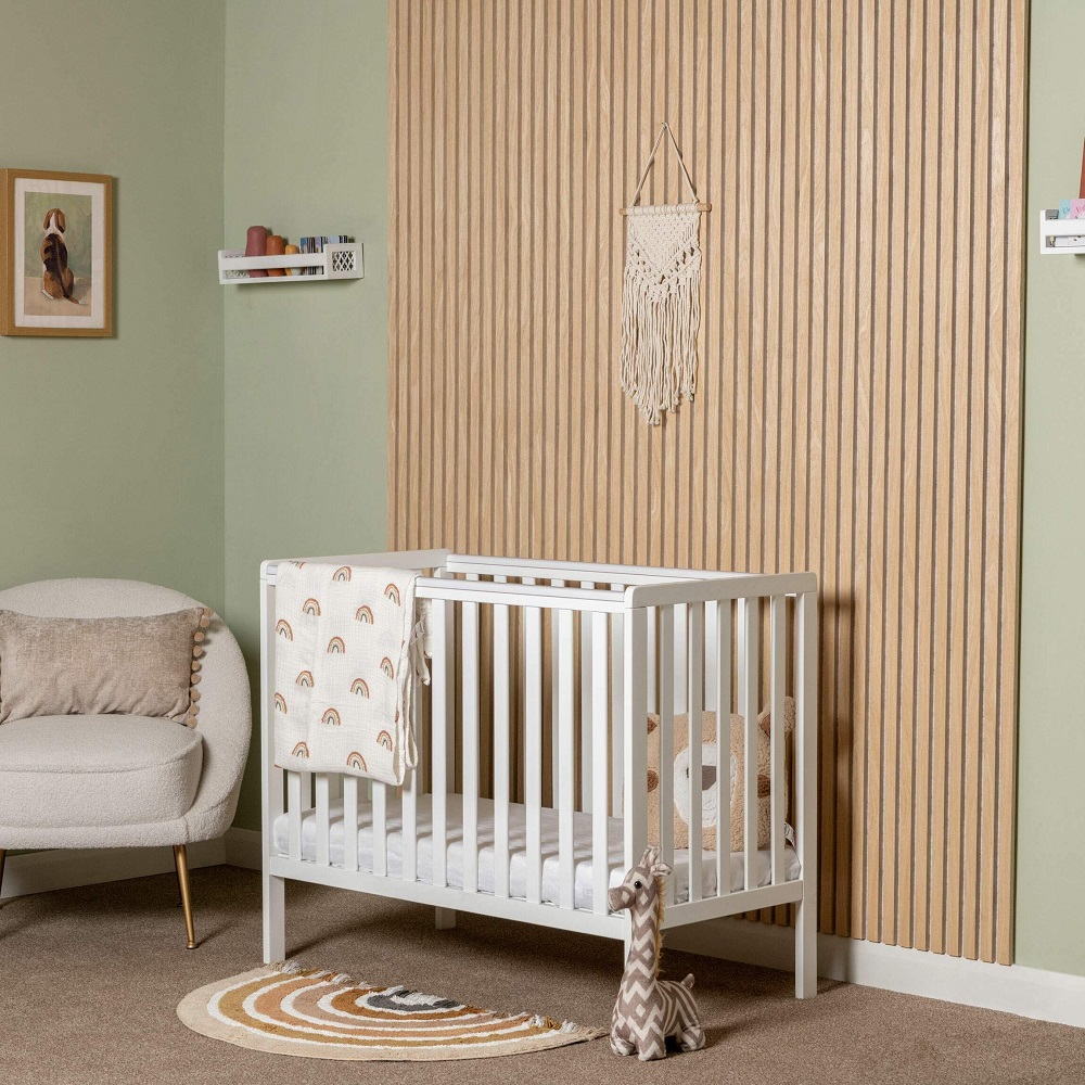 Warm and inviting nursery room with oak acoustic wall slat panels and grey felt, featuring a white crib with a rainbow patterned blanket, a comfortable armchair, and a playful giraffe toy on a rainbow shaped rug, all under a macrame wall hanging. 