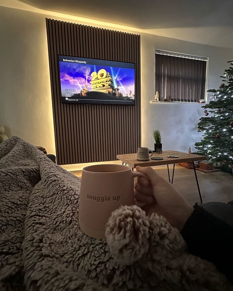 Cosy living room ambiance with a person holding a 'snuggle up' mug, enjoying a relaxing moment on a sofa with a soft, fluffy blanket. In the backgrounf, a wall mounted TV is set against a warm wooden slat wall feature, displaying a vibrant home entertainment screen. The space is further enhanced by a subtle holiday vibe with a Christmas tree and seasonal window decorations, inviting comfort and festive cheer into the home interior design. 