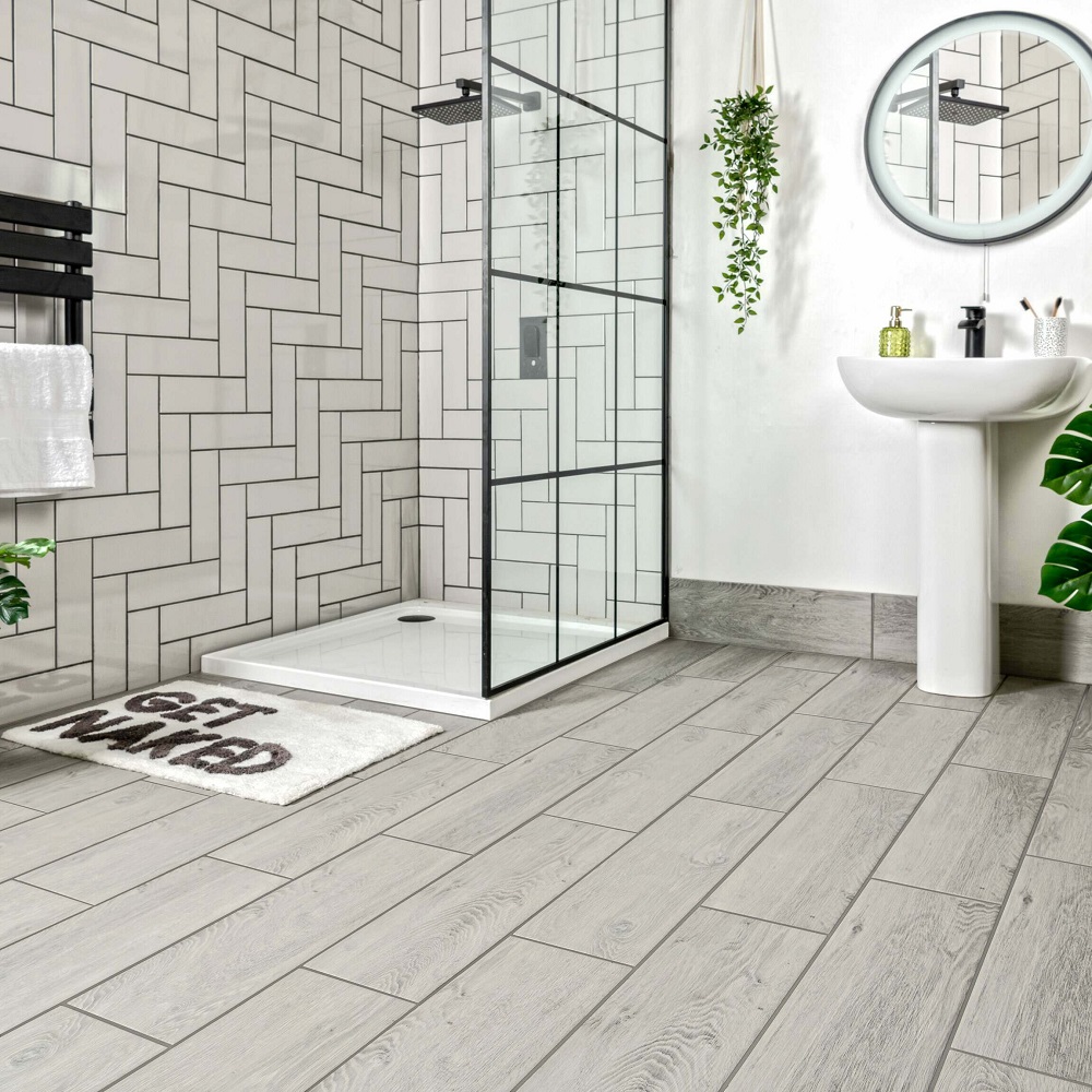 Elegant grey wood effect floor tiles in a spacious bathroom with chic geometric white wall tiles, sleek glass shower enclosure and fresh greenery enhancing the tranquil spa-like atmosphere. 