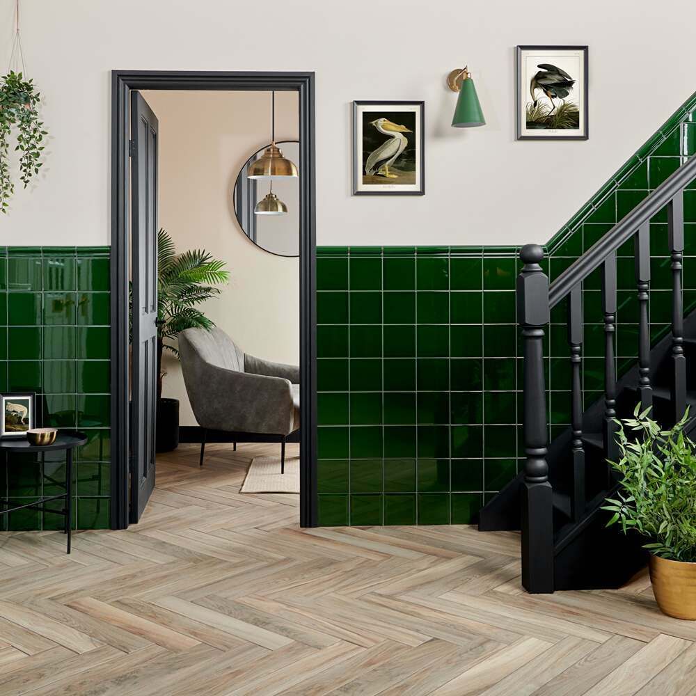 A vibrant and inviting hallway that pairs rich Victorian green subway tiles with a herringbone parquet floor, creating a harmonious blend of classic and contemporary design elements. The glossy green tiles rise to dado height, adding a pop of colour and easy to clean practicality. A sleek black staircase with matchinf spindles anchors the space, leading to an upper level. Natural light reflects off a round mirror, complementing the open door which reveals a glimpse of a cosy sitting area. Artwork featuring bird illustrations and a wall mounted green sconce add character, while indoor plants introduce a refreshing touch of greenery. 