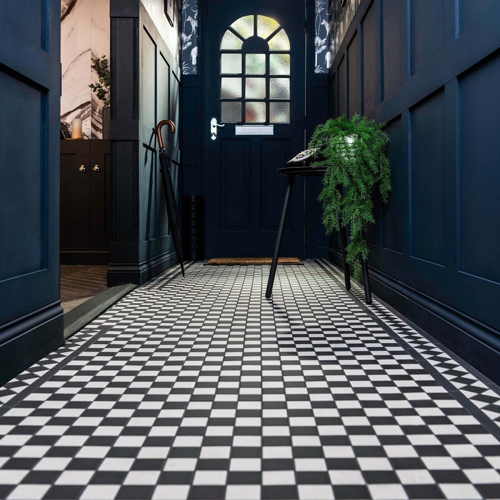 This hallway exudes classic sophistication with its Churchill Snow & Midnight Chequer mosaic tiles. The high-contrast black and white chequered pattern creates a dynamic, eye-catching floor that pairs beautifully with the deep navy panelled walls. A stained glass door adds a vintage touch, while the understated elegance of the dark wood side table and the lush green of a potted fern plant breathe life into this space. The scene is complete with traditional elements like a wooden can and a woven mat, enhancing the welcoming ambiance. 