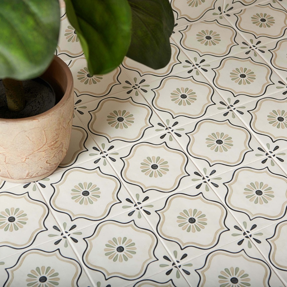 A close up view of decorative floor tiles with a floral pattern, featuring a colour palette of sandy beige, olive green and white. A potted plant with large green leaves partially obscures the view on the top left corner. 