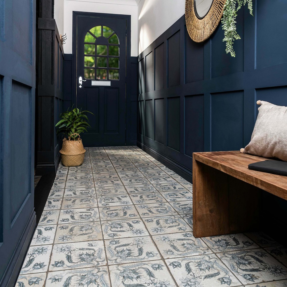An elegant hallway breathes sophistication with patterned tiles, their vintage motifs in indigo blue and ivory creating an ornate tapestry underfoot. This grand entrance is framed by walls and a door painted in a deep, navy blue, with wainscoting adding architectural detail. A natural wood bench offers a place to rest, while plush greenery in a woven basket brings a touch of nature indoors. A round mirror adorned with a rustic charm hangs above, reflecting the stately space and amplifying the natural light the filters through the arched window on the door. 