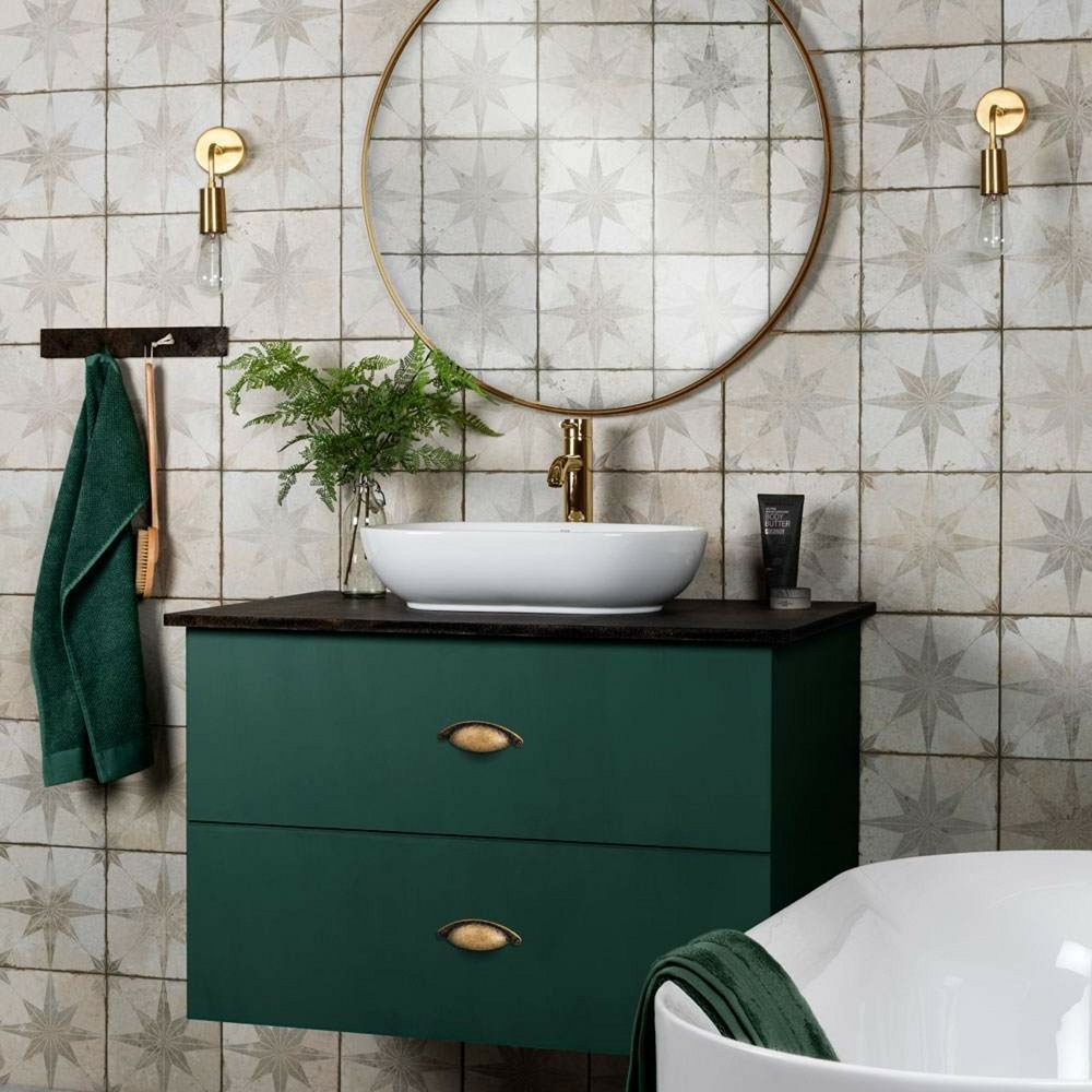 A chic bathroom vignette with a deep forest green vanity unit, topped with a contrasting white vessel sink and adorned with brass fixtures. The vanity is fitted with two drawers, each featuring vintage brass  wall sconces with exposed bulbs. The walls are tiled with Sintilla Dusk star-patterned tiles, adding a subtle vintage charm to the space. A lush green hand towel hangs to the side, echoing the vanity's colour, and a small potted fern adds a fresh touch of greenery. 