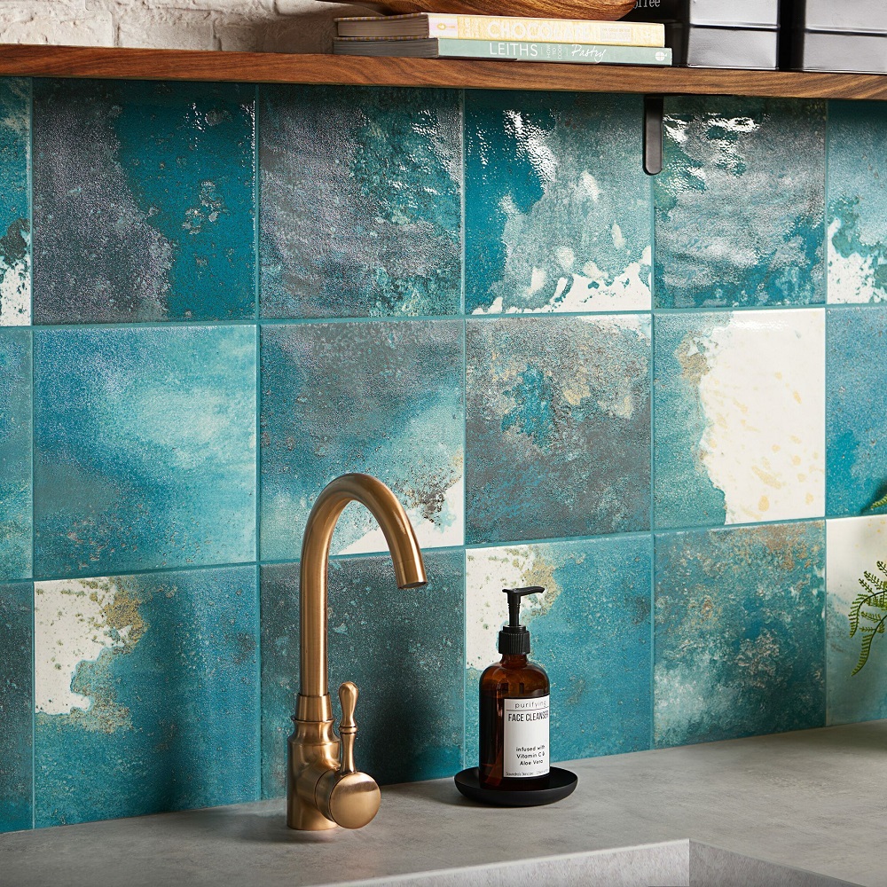 A close-up of a kitchen backsplash featuring Raku square tiles with a unique design, presenting shades of turquoise and weathered copper effects. The tiles are complemented by a sleek countertop and a brass faucet with a classic design. Resting beside the faucet is an amber glass bottle of face cleanser with a black pump, placed on a dark round tray. Above the splashback, a wooden shelf holds an assortment of cookbooks, adding a personal and homey touch to the modern, rustic aesthetic of the space. 