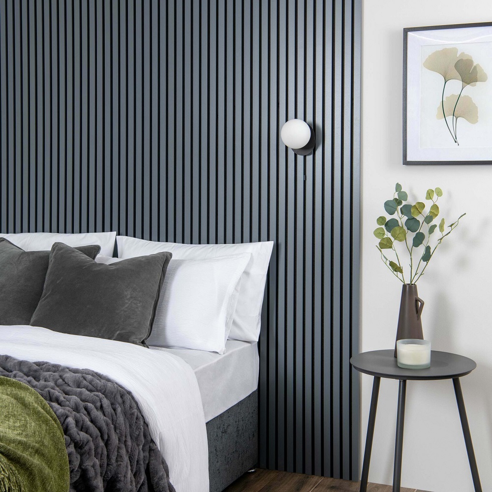 A modern bedroom featuring a wall with vertical, carbon-coloured acoustic wood panels. The bed has crisp white bedding, complemented by dark grey pillows and a textured throw. A sleek bedside table holds a simple white spherical lamp and a tall vase with eucalyptus branches, next to a framed botanical print. 