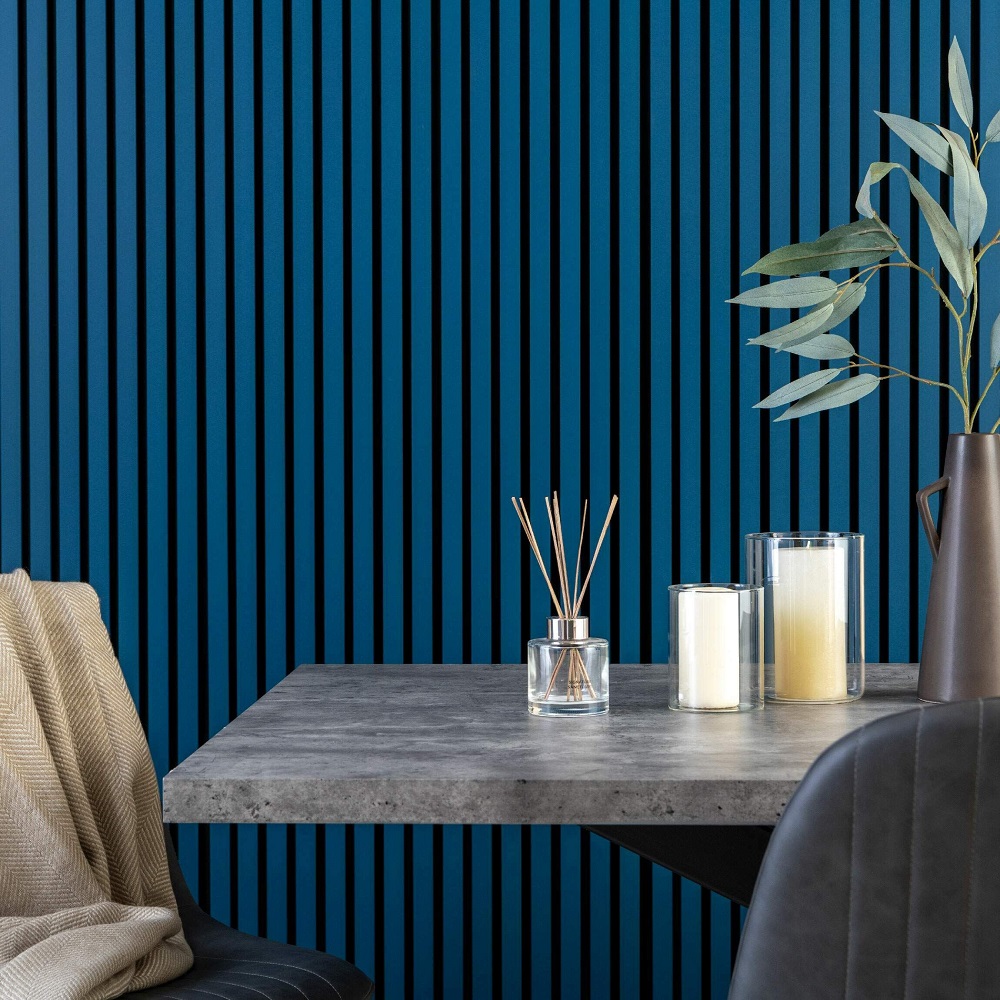 A contemporary dining space showcasing a bold feature wall with vertical acoustic wood panels in a striking denim blue colour, contrasted by black felt backing to act as wallpaper alternative. A concrete style dining table adds an industrial touch, complemented by a cosy cream throw draped over the chair in the foreground. The table is adorned with a minimalist arrangement including a reed diffuser in a clear glass bottle, and two off white candles in varying heights. A tall vase with eucalyptus branches lends a touch of organic warmth to the chic, modern composition. 