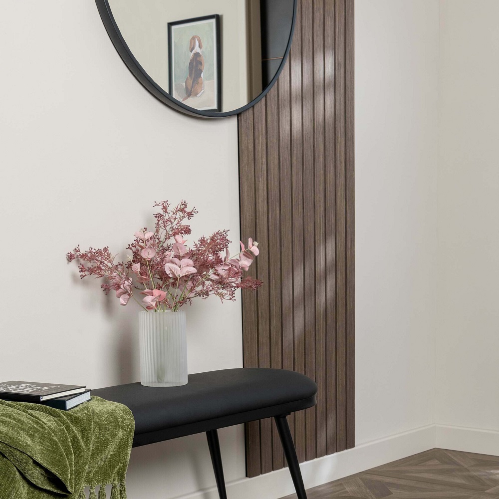 A serene and modern hallway corner characterised by it acoustic slat panelling, providing a warm and textured backdrop. The space is thoughtfully accented with a sleek black bench, on which rests an elegant glass vase holding delicate pink blossoms that add a splash of colour. A round black framed mirror reflects the simple yet striking vignette, enhancing the sense of space, while a plush green throw adds a hint of cosiness. The setting is grounded by herringbone parquet flooring that complements the walnut tones, creating a tranquil entryway retreat. 