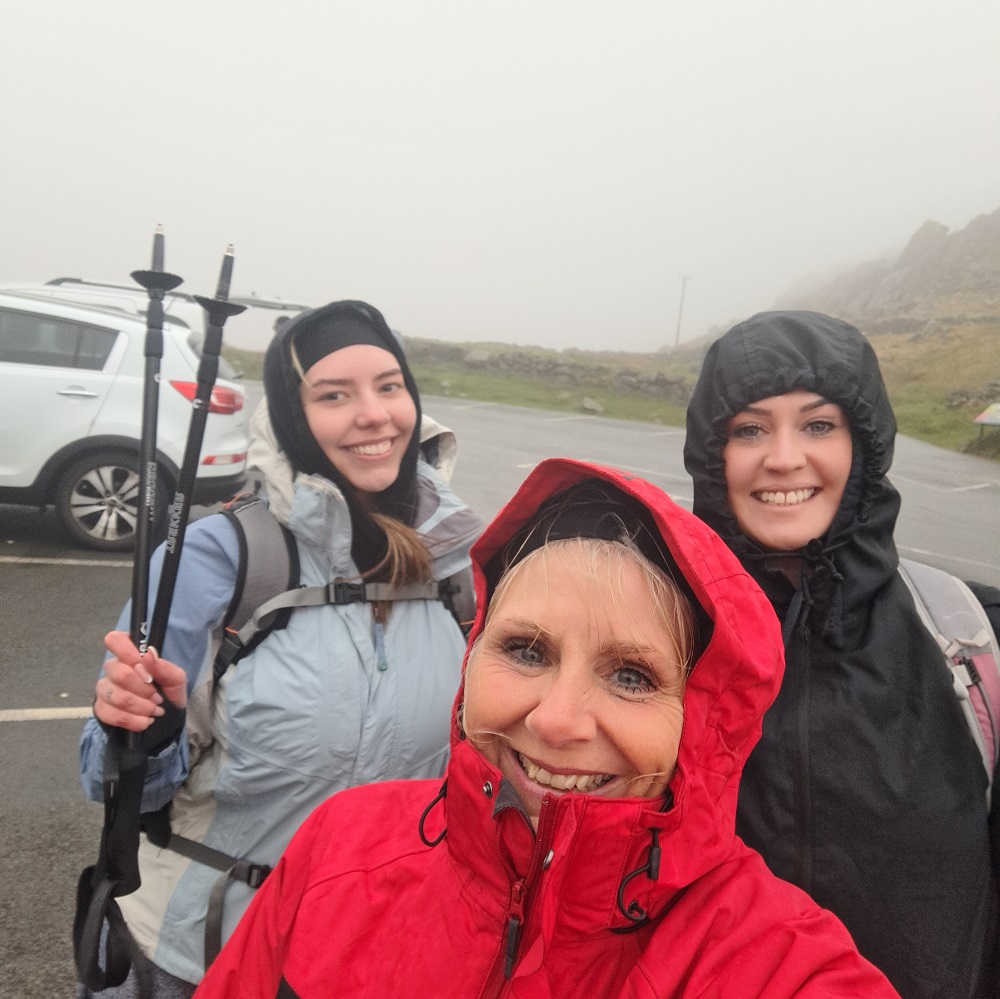 Three cheerful hikers posing for a selfie on a foggy day: three women in selfie, one in a red raincoat, one in a blue jacket and the other in a black raincoat. 