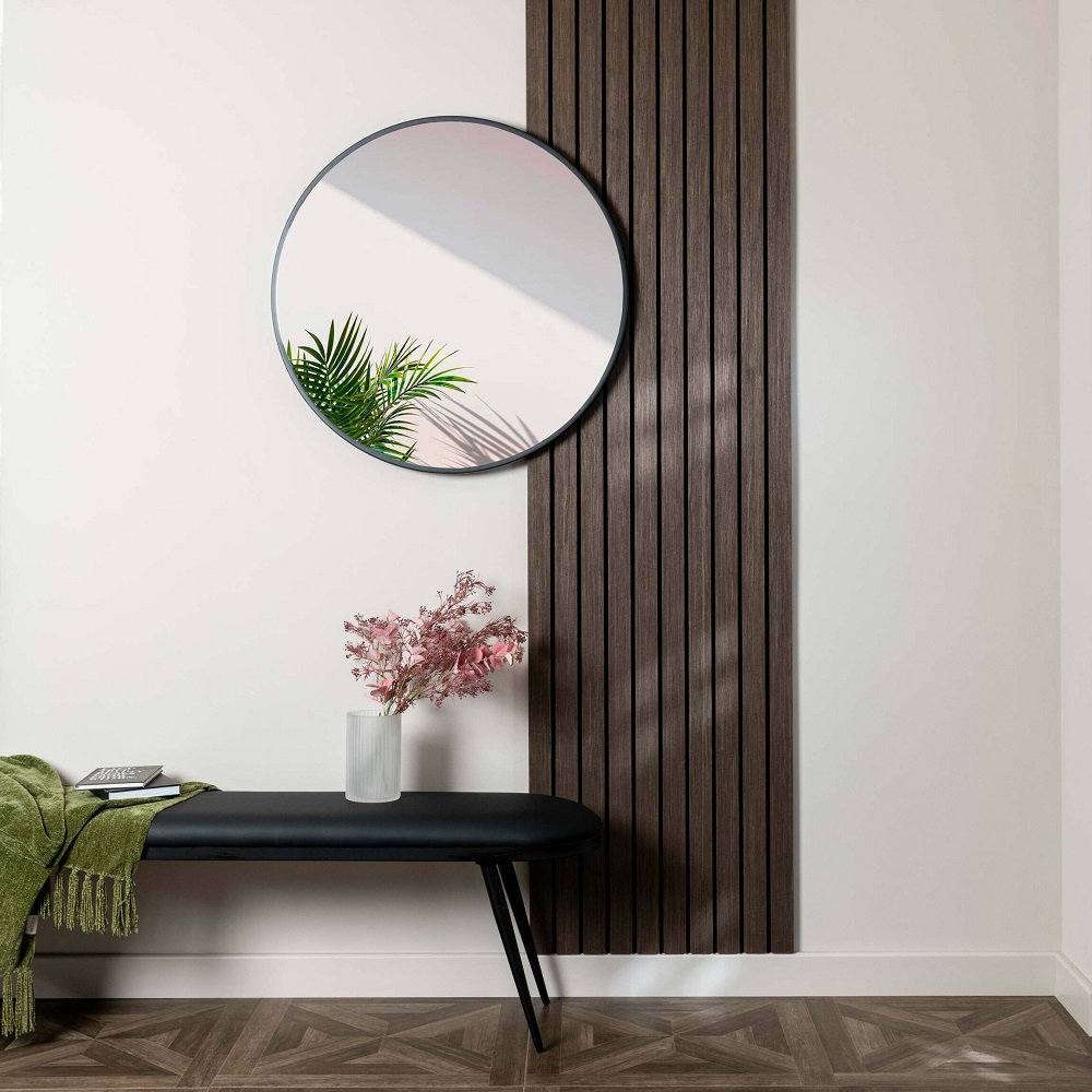 A minimalist hallway featuring a walnut wide acoustic wood panel on one side, paired with a simple white wall. A large round mirror reflects a hint of greenery, enhancing the space. Below, a sleek black bench with a pink floral arrangement in a white vase and a green throw blanket invites a welcoming atmosphere. The herringbone-patterned wooden floor adds warmth and texture to the space. 